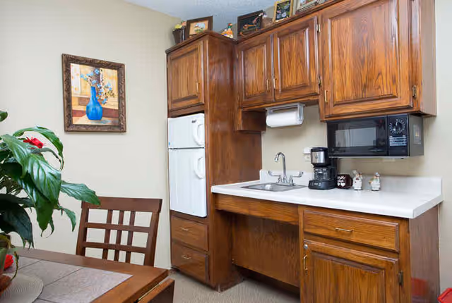 A small kitchen area with wooden cabinets, a white countertop, a sink, a coffee maker, a microwave, and a refrigerator. There is a wooden dining table with chairs and a potted plant with red flowers. A framed painting of a blue vase with flowers hangs on the wall.