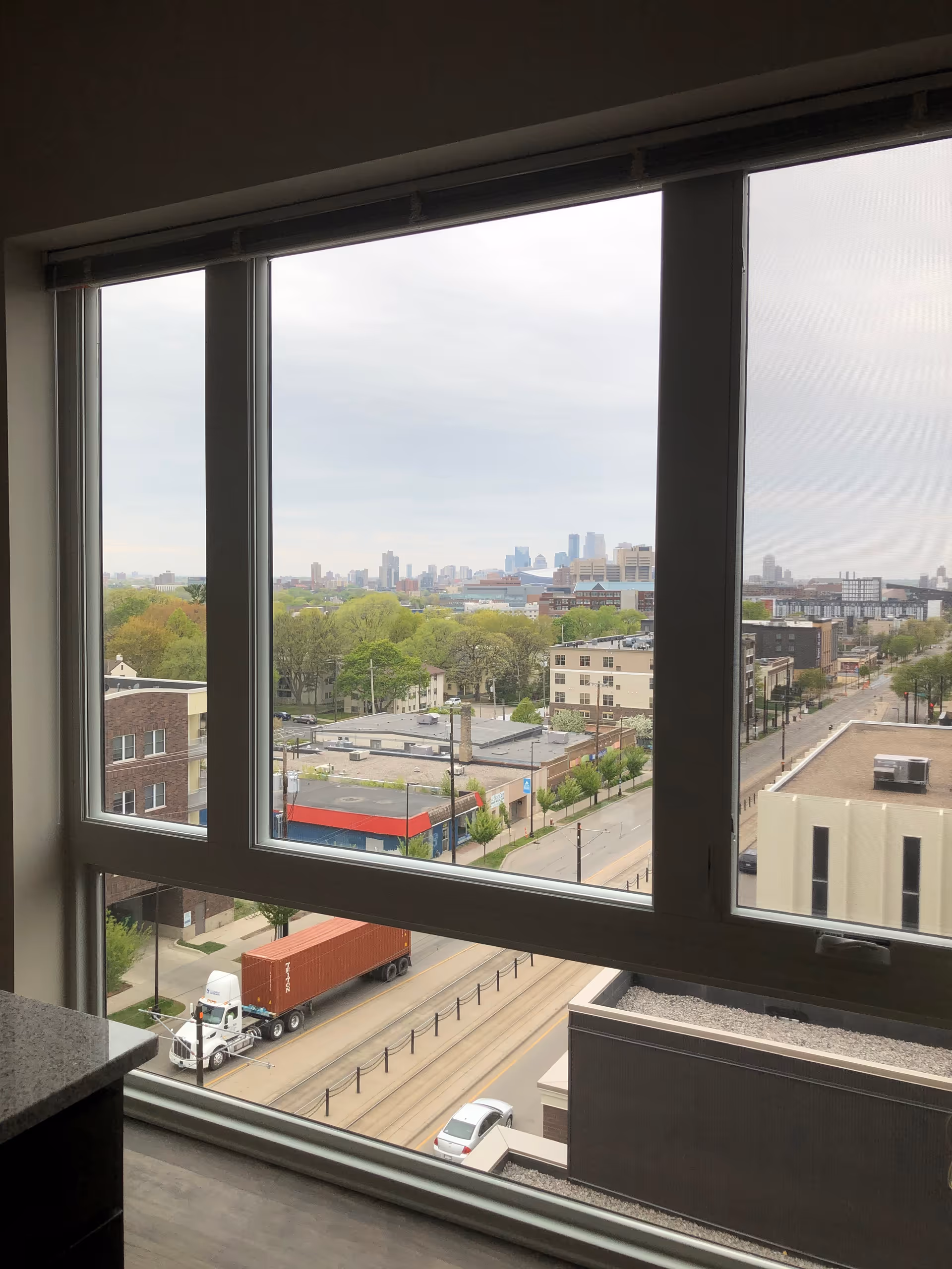 View through a large apartment window overlooking a city street with a parked truck, nearby buildings, trees, and a distant skyline.