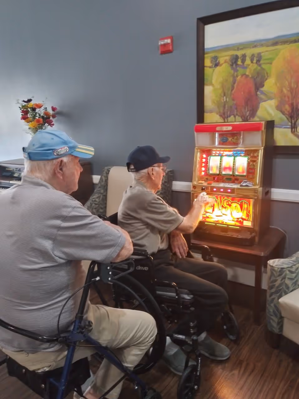 Two elderly men sitting in wheelchairs playing a slot machine in a room with a painting of a landscape on the wall and a vase of colorful flowers on a table nearby.