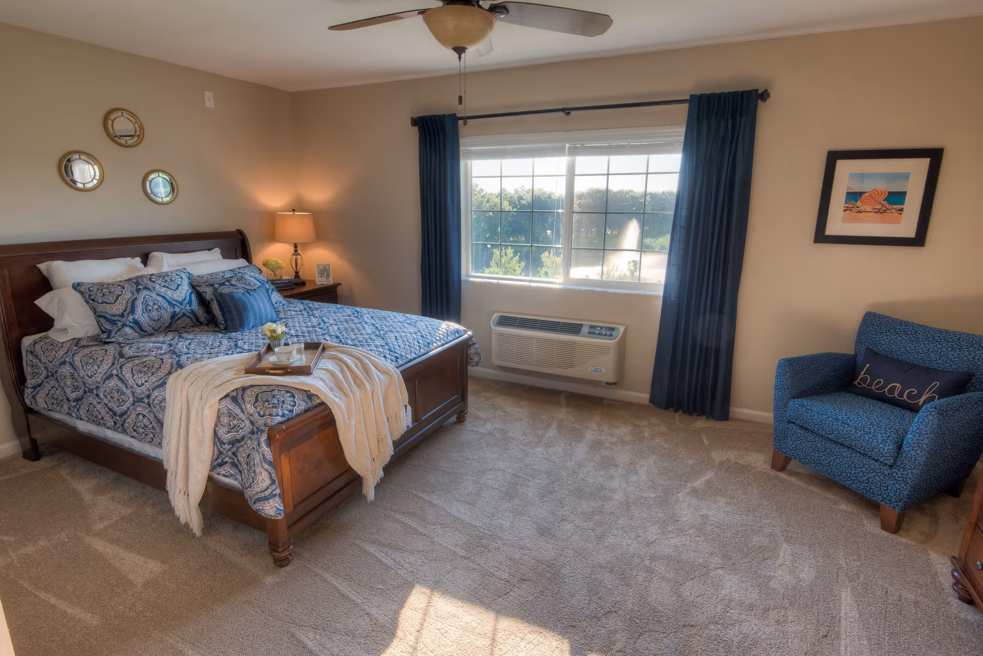 A cozy bedroom with a wooden bed frame, blue patterned bedding, and several pillows. A beige throw blanket is draped over the foot of the bed with a tray holding a small flower vase and a glass. There is a nightstand with a lamp and a small plant next to the bed. The room has beige carpet, a ceiling fan, a large window with blue curtains, and a blue armchair with a pillow that says 'beach'. Three round decorative mirrors hang on the wall above the bed.