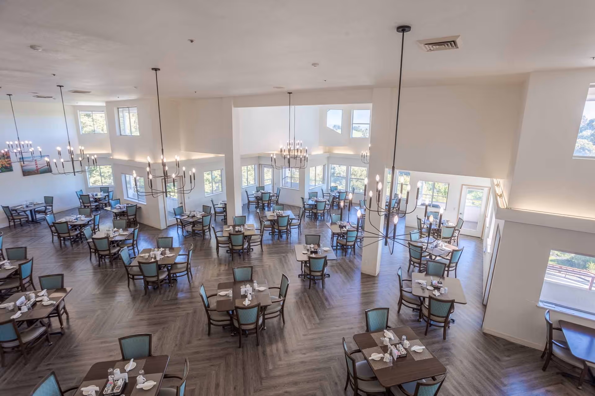 Spacious dining room with multiple square tables set with white napkins and utensils, surrounded by chairs with teal upholstery. The room has large windows allowing natural light to fill the space, modern chandeliers hanging from the ceiling, and light-colored walls with wood-patterned flooring.