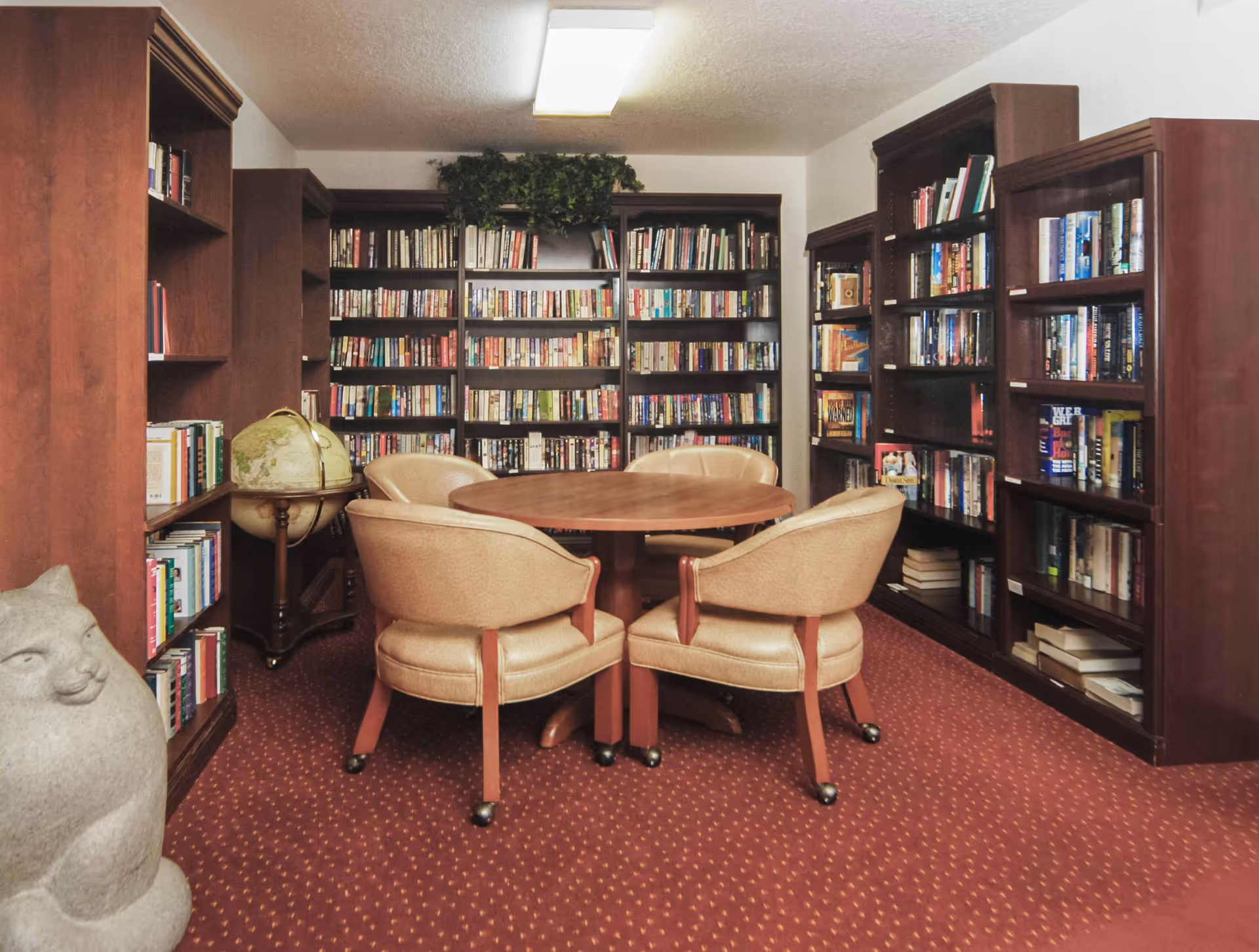A cozy library room with wooden bookshelves filled with books lining the walls. In the center, there is a round wooden table surrounded by four beige cushioned chairs on wheels. A globe on a stand is positioned to the left, and a stone sculpture resembling a cat is visible in the foreground on the left side. The room has a red carpet with a subtle pattern and a ceiling light fixture.