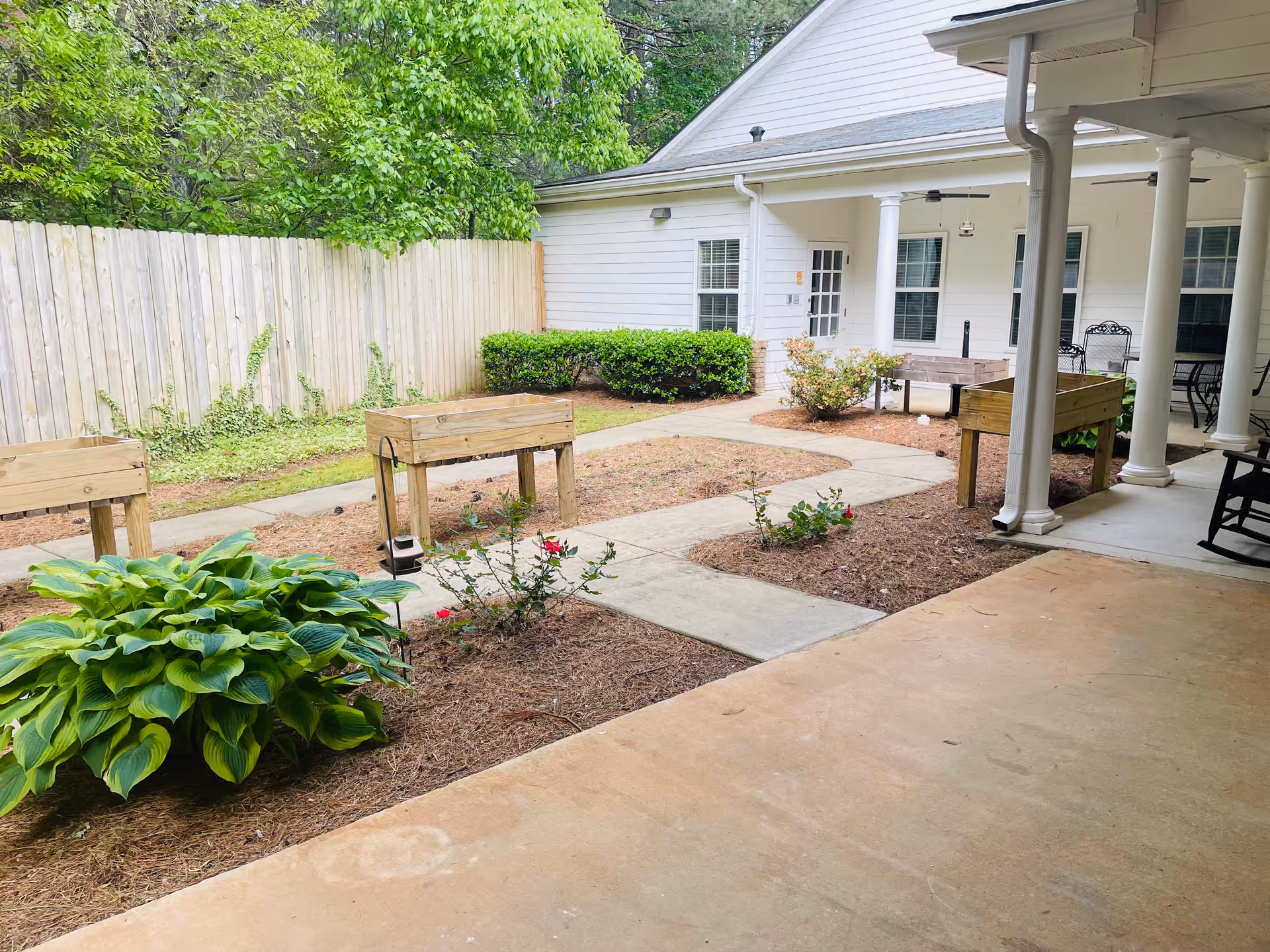 Outdoor courtyard area with raised wooden garden beds, small plants, and a concrete walkway. The area is surrounded by a wooden fence and greenery, with a white building featuring windows and a covered patio with columns and outdoor furniture.