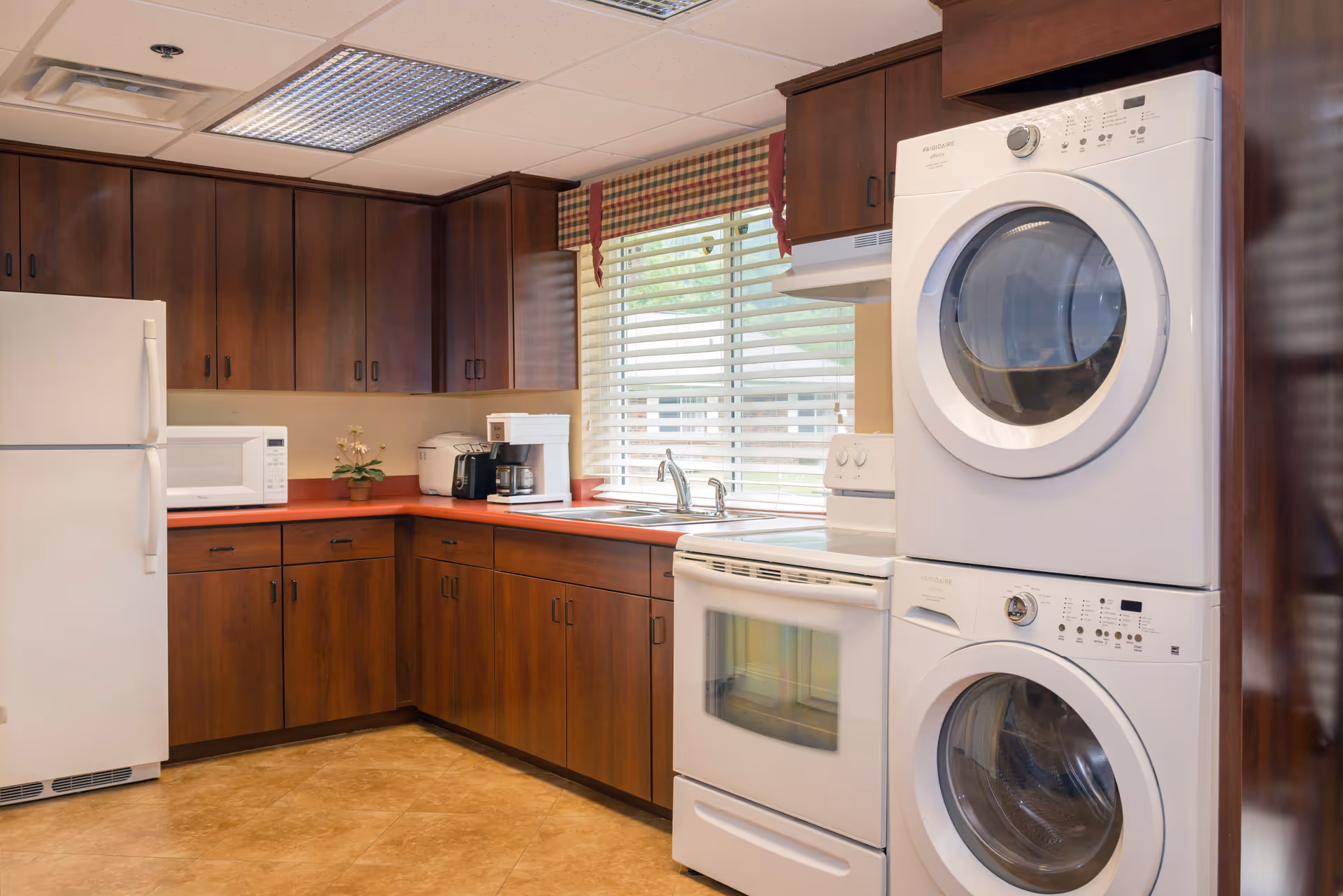 A kitchen area with dark wooden cabinets, a white refrigerator, microwave, coffee maker, and a stacked washer and dryer unit. There is a window with blinds and a plaid valance above the sink.