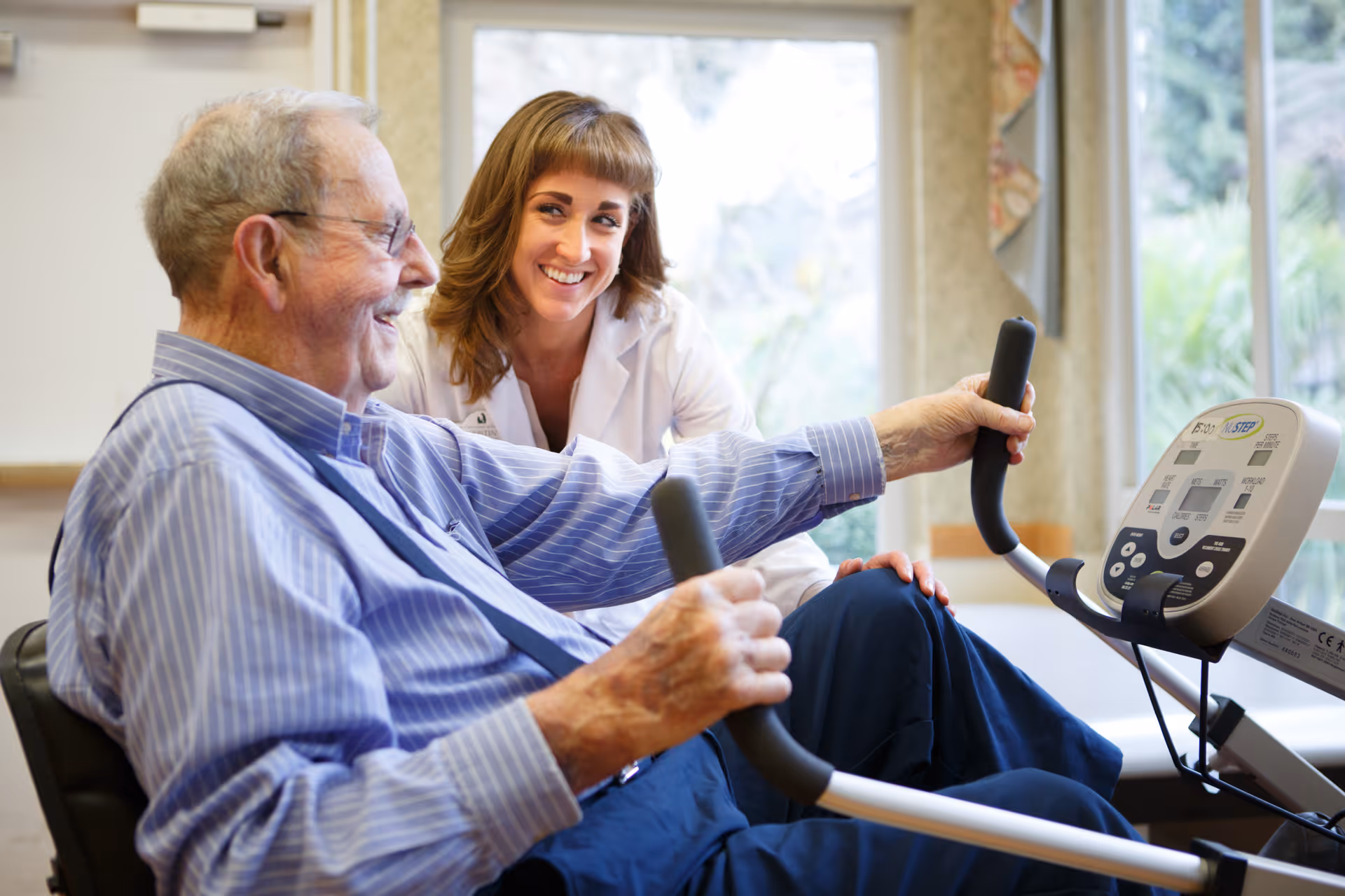 An elderly man using a seated exercise machine while a female healthcare professional in a white coat smiles and supports him in a bright room with large windows.