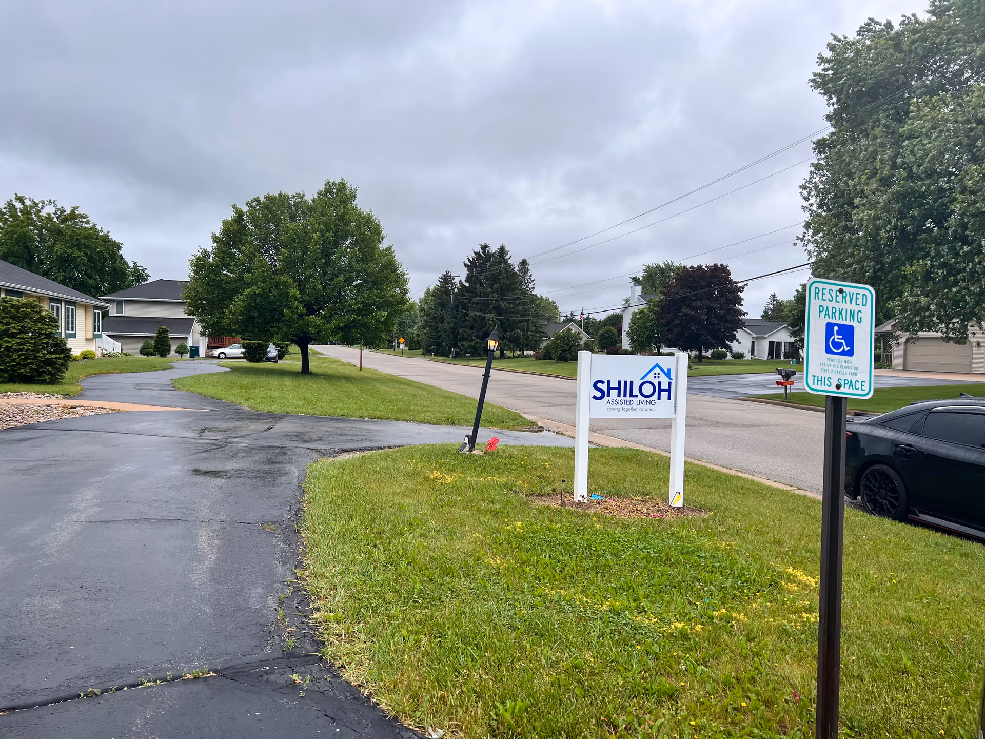 Street view showing a Shiloh Assisted Living sign on a grassy lawn with a reserved handicapped parking sign, nearby houses, and a cloudy sky.