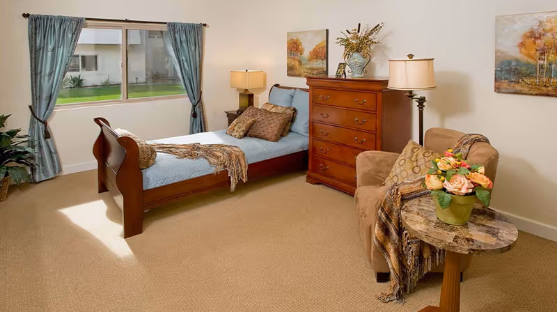 A cozy bedroom with a wooden bed featuring blue bedding and multiple pillows, a wooden dresser with a lamp and decorative items on top, a comfortable armchair with a throw blanket and pillow, a small round table with a flower arrangement, and two paintings on the walls. A window with blue curtains lets in natural light and shows a green lawn outside.