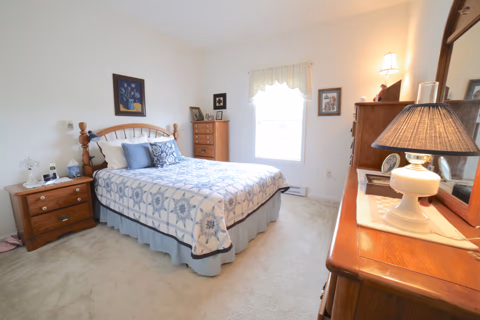 A cozy bedroom with a wooden bed frame and a bed covered with a blue and white quilt. There are multiple pillows on the bed. To the left of the bed is a wooden nightstand with a lamp, phone, and other small items. A wooden dresser with a mirror and a lamp is on the right side of the room. The room has a window with a white curtain, light-colored carpet, and framed pictures on the walls.