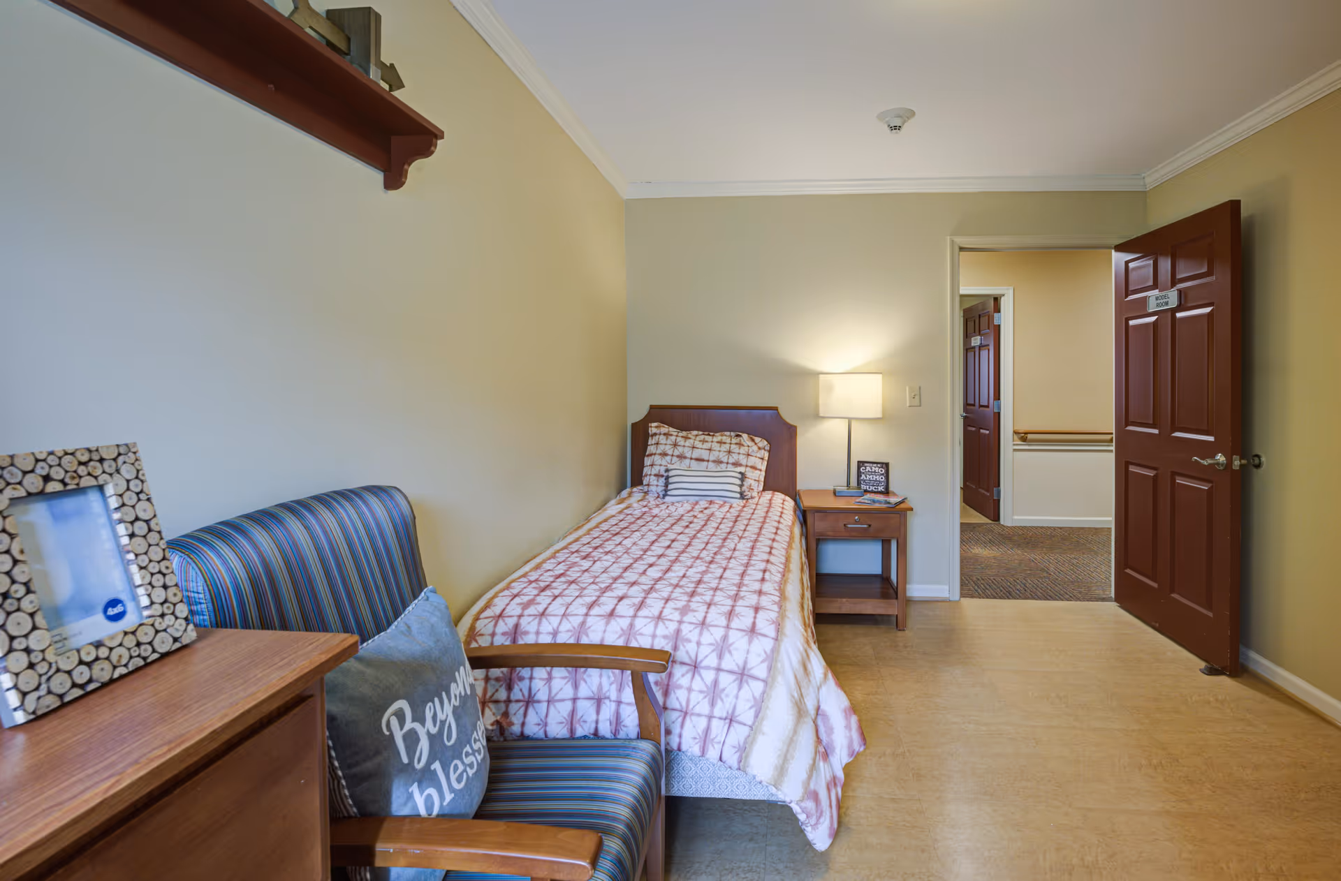 A small bedroom with a single bed covered with a patterned quilt, a wooden nightstand with a lamp and a book, a striped armchair with a pillow that says 'Beyond blessed', and a wooden dresser with a photo frame. The door to the room is open, showing a hallway outside.