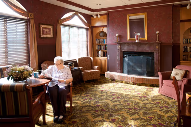 An elderly woman sitting at a wooden table reading a newspaper in a cozy living room with patterned carpet, upholstered chairs, a fireplace with a mirror above it, bookshelves, and large windows with blinds and curtains letting in sunlight.
