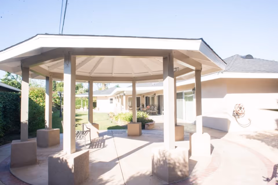Outdoor gazebo structure with a white roof and beige pillars in a courtyard area of a senior living facility. Surrounding the gazebo are green bushes, a lawn, and a beige building with sliding glass doors and a garden hose mounted on the wall.