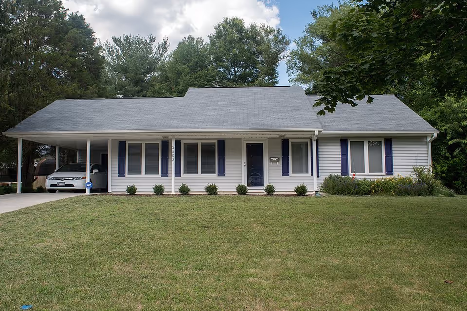 Single-story assisted living facility front with carport, blue shutters, and a manicured lawn.