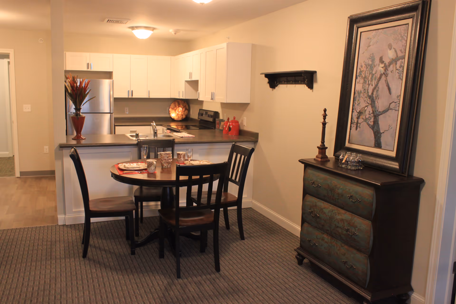 A dining area with a round wooden table set for four with plates, cups, and glasses. Behind the table is a kitchen with white cabinets, a stainless steel refrigerator, and a countertop with a sink. To the right, there is a decorative chest of drawers with a framed painting of birds on a tree above it. The room has beige walls and carpeted flooring.