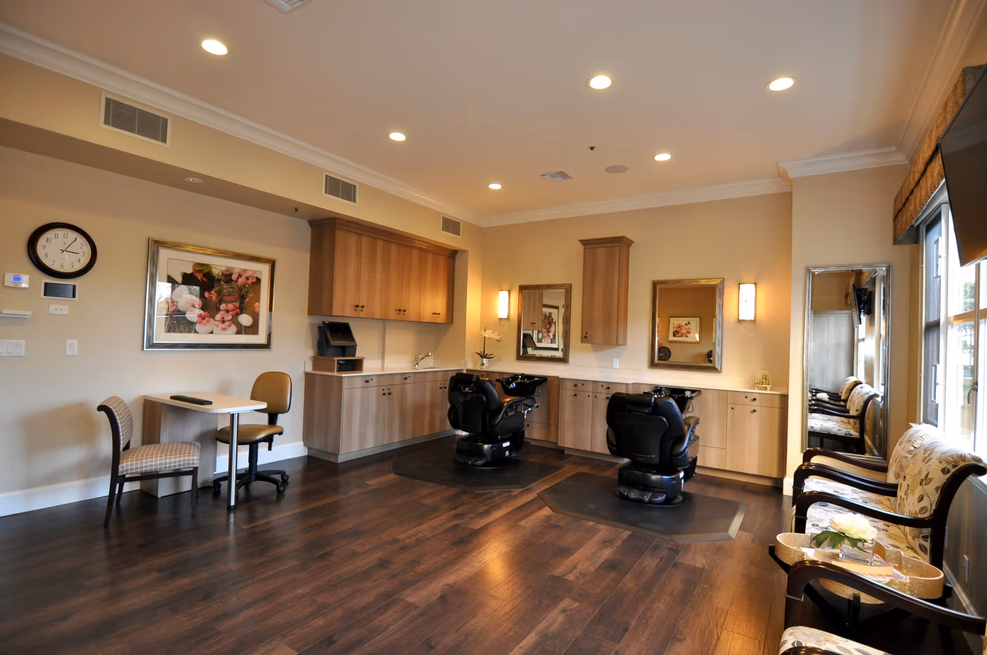 A well-lit salon room with two black salon chairs in front of mirrors mounted on a beige wall with wooden cabinets underneath. The room has dark wooden flooring, a small table with two chairs on the left side, and a seating area with cushioned chairs and a small table with flowers near a large window on the right side.