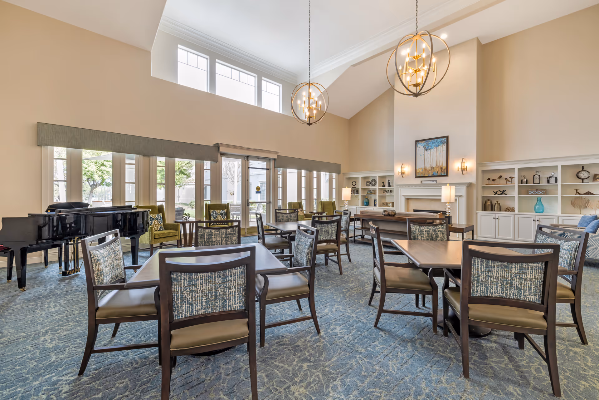 A spacious and well-lit common area with multiple tables and chairs arranged on a patterned carpet. The room features large windows allowing natural light to enter, a black grand piano on the left, green armchairs near the windows, and built-in white shelves with decorative items. Two modern spherical chandeliers hang from the high ceiling, and a fireplace with a painting above it is centered on the far wall.