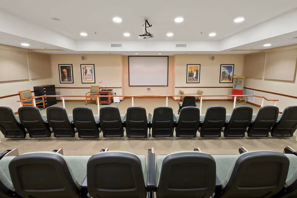 A small auditorium or presentation room with two rows of black cushioned chairs facing a blank projection screen on the front wall. The room has beige walls with framed posters, recessed ceiling lights, and a popcorn machine in the corner. There are also a few wooden chairs and a small table near the front.