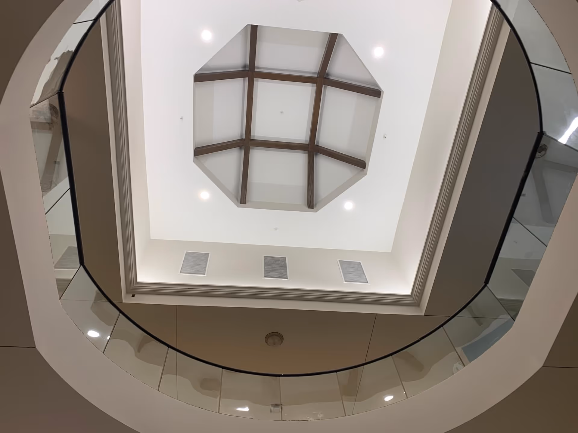View looking up at a modern ceiling with an octagonal skylight framed by wooden beams, surrounded by recessed lighting and a circular glass railing below.