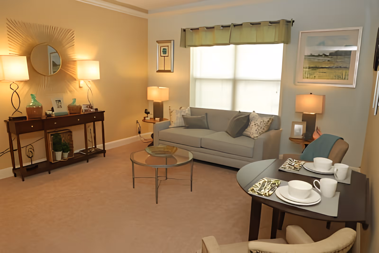 A cozy living room with a gray sofa adorned with pillows, a glass coffee table, and a small dining table set for two with white dishes and cups. The room has beige carpet, warm lighting from table lamps, a decorative mirror on the wall, and framed artwork near a window with a green valance.