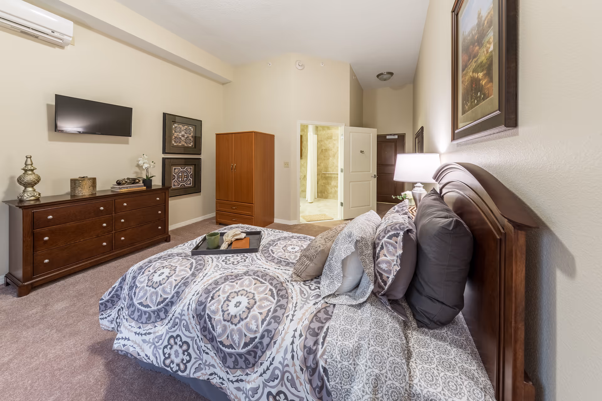 Bedroom with patterned bedding, wooden headboard, dresser with a wall-mounted TV, wardrobe, and an open door leading to a bathroom.