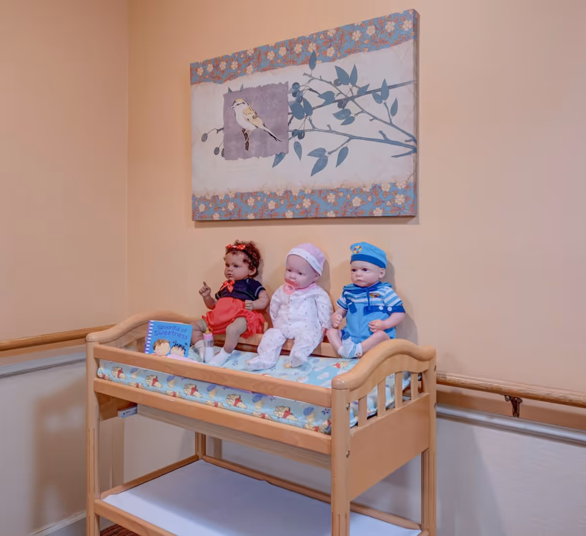 A wooden changing table with a padded surface featuring a cartoon pattern, holding three baby dolls dressed in different outfits. Behind the dolls is a book titled 'Spoonful of Sweetness.' Above the table hangs a framed artwork of a bird perched on a branch with a floral border. The wall is painted a soft peach color with a wooden handrail running horizontally along it.