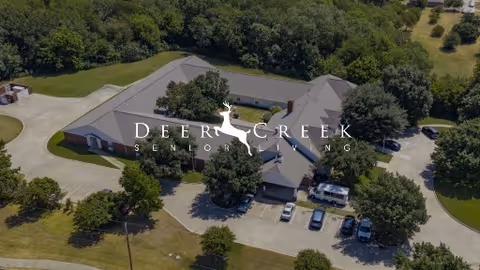 Aerial view of a single-story senior living building with driveways, parked cars and surrounding trees, with a Deer Creek Senior Living logo overlaid.