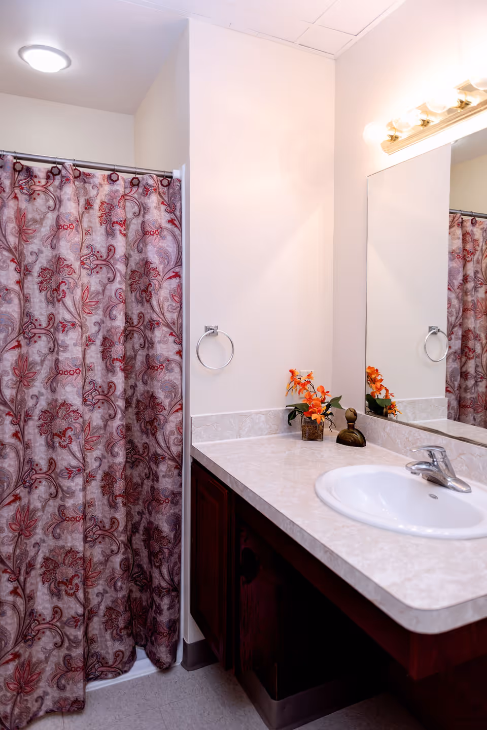 A bathroom with a white sink set in a marble countertop, a large mirror above the sink, a towel ring on the wall, and a shower with a decorative red and beige patterned curtain. There is a small vase with orange flowers on the countertop.