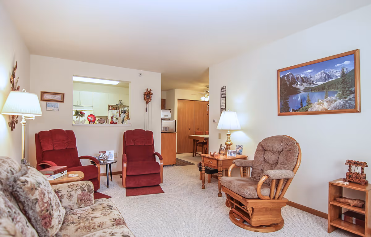 Furnished living room in a senior apartment featuring red recliners, a floral sofa, a wooden rocking chair, side tables, and a mountain landscape painting.