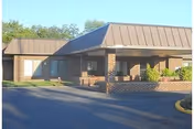Exterior view of a single-story brick building with a covered entrance and a metal roof, surrounded by a paved driveway and some greenery in the background.