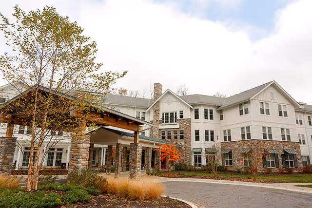 Front entrance of a multi-story senior living building with a stone-columned porte-cochere, landscaped grounds, and a circular driveway.