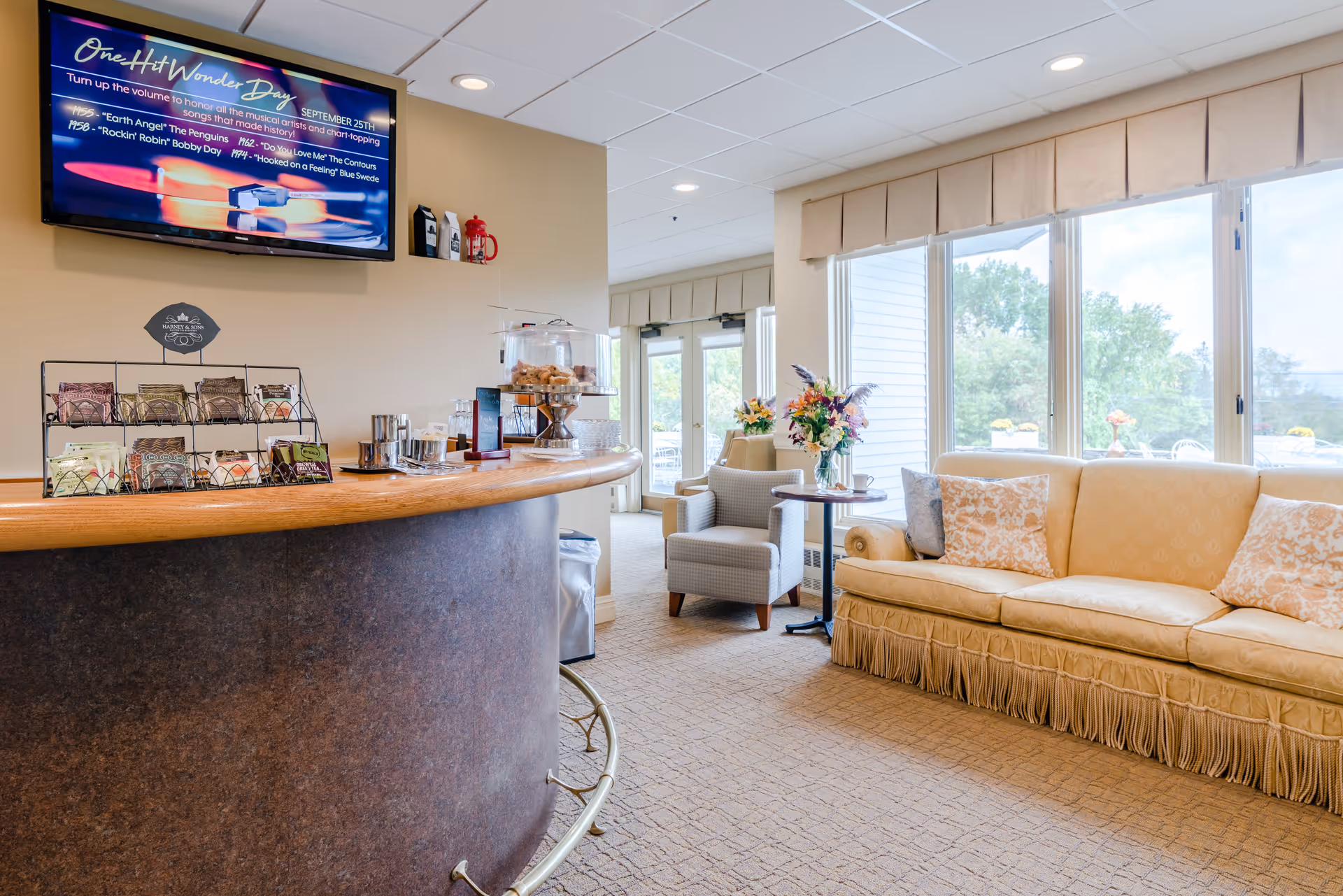 Bright senior living lobby with a curved reception counter, seating area with sofa and armchairs, and large windows overlooking greenery.