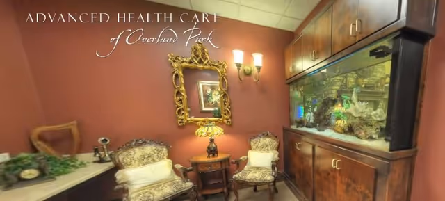 A cozy sitting area with two ornate upholstered chairs with white cushions, a small wooden side table with a decorative lamp, a large built-in aquarium filled with coral and fish, a gold-framed mirror on the wall, and warm lighting fixtures on a reddish-brown wall.