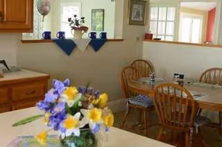 A cozy dining area with a wooden table and chairs, set with placemats and condiments. In the foreground, there is a vase with colorful flowers including yellow daffodils and purple blooms. The background shows a half wall with a shelf holding blue mugs and a potted plant, and a kitchen area with wooden cabinets.