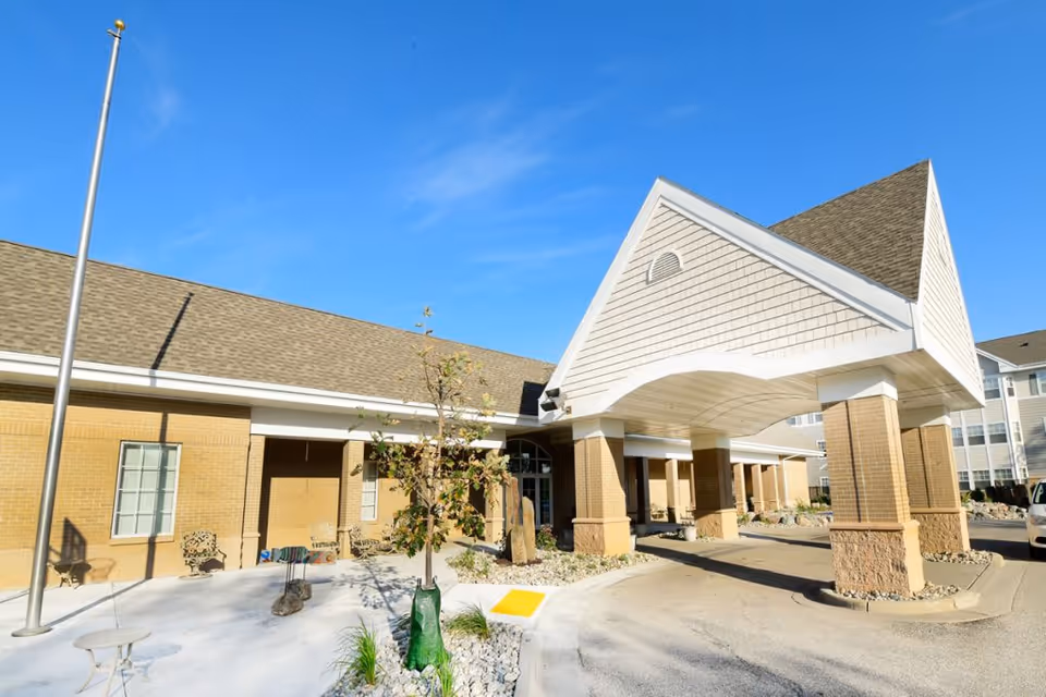 Front entrance and covered porte-cochère of a senior living facility with brick columns, a flagpole, and a clear blue sky.