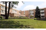 Exterior view of a multi-story brick retirement community building with several windows, surrounded by green grass, trees, and shrubs under a partly cloudy sky.