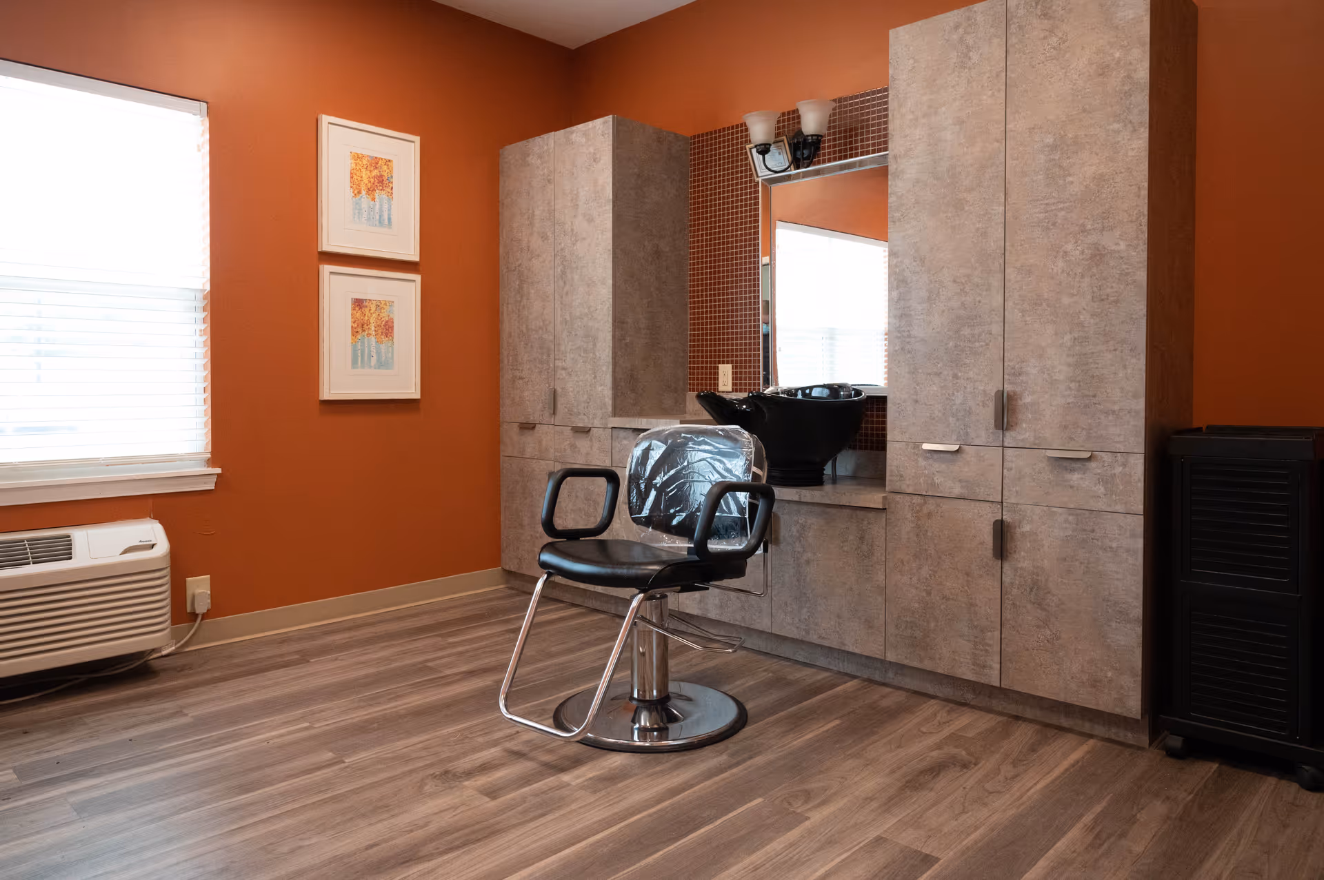 Interior view of a salon area with a black salon chair in front of a countertop with a black wash basin. The walls are painted orange, and there are two framed pictures on the wall next to a window with blinds. The floor is wood laminate, and there are large cabinets for storage.