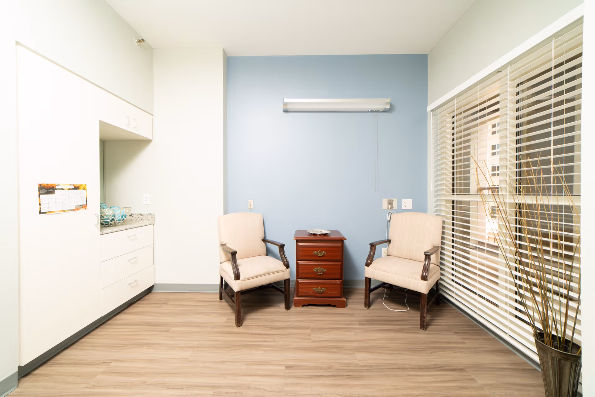A small sitting area in a healthcare facility with two beige armchairs placed on either side of a wooden three-drawer nightstand against a light blue wall. To the left, there is a white cabinet with drawers and a granite countertop. On the right, there is a large window with white blinds and a tall vase with decorative sticks. The floor is wood-patterned.