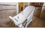 A large white therapeutic bathtub with handrails and jets, situated in a tiled room with beige and brown tiles on the walls and floor. The room appears to be designed for assisted bathing, likely in a nursing or care facility.