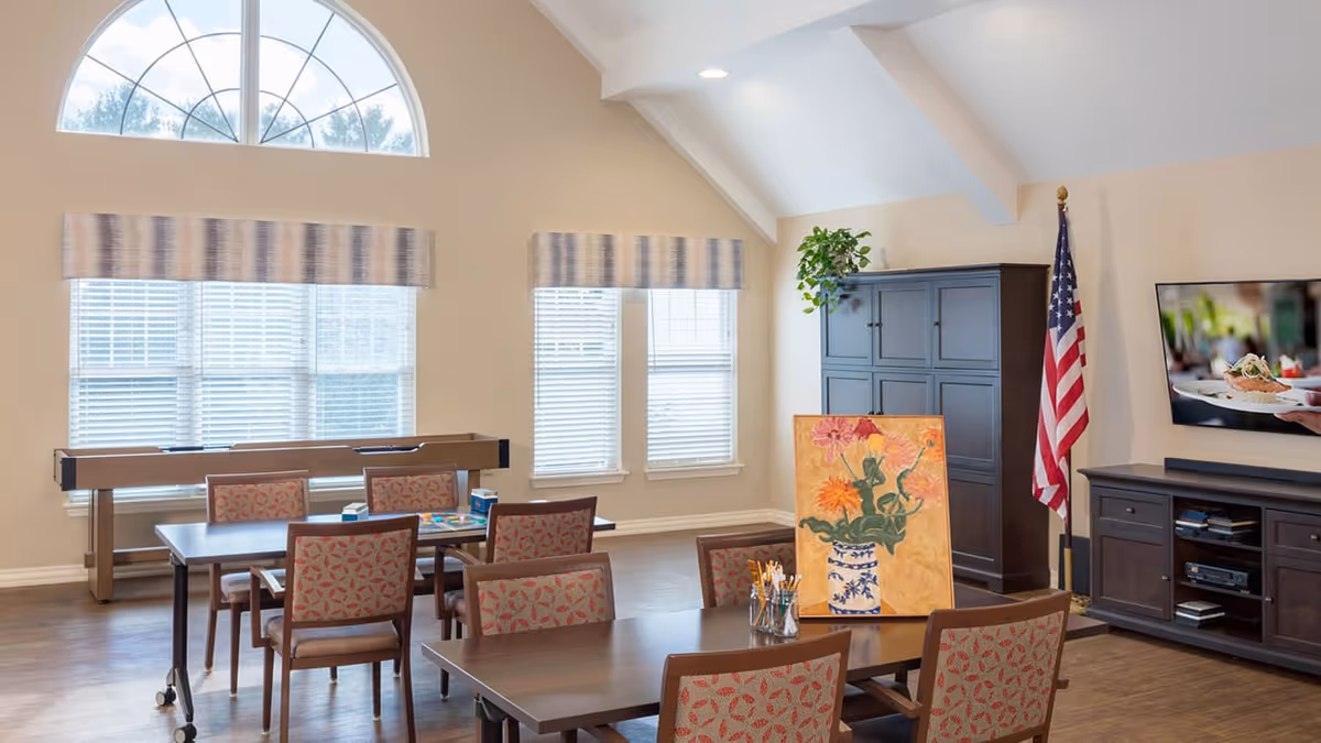A bright room with high vaulted ceilings and large windows with blinds and valances. There are two tables with patterned chairs around them, one table has art supplies and a painting of flowers in a vase. In the background, there is a foosball table, a tall dark cabinet with a plant on top, an American flag, and a wall-mounted TV showing a close-up of a plated meal.