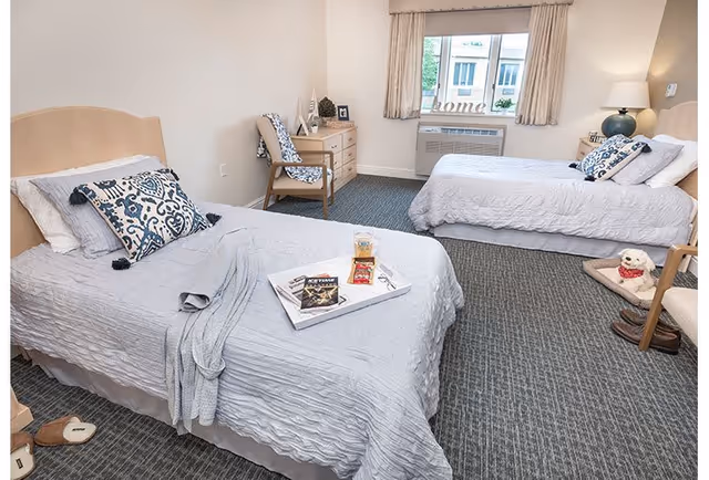 A bright senior living bedroom with two twin beds covered in light gray bedding and decorative pillows. A tray with snacks and magazines is placed on one bed. The room has a window with beige curtains, a wooden dresser, a chair with a patterned cushion, a lamp on a side table, and a small dog bed with a stuffed dog toy. The carpet is gray and textured.