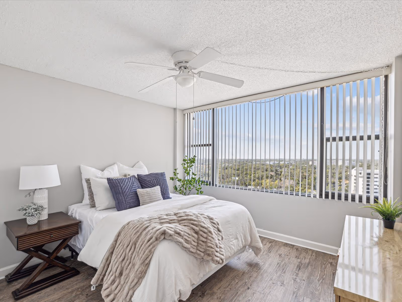 A bright bedroom with a large window covered by vertical blinds showing a view of trees and buildings outside. The room features a bed with white bedding, several pillows in white and purple, and a beige textured throw blanket. There is a wooden nightstand with a white lamp and a small plant, a ceiling fan with light, and a wooden dresser with a small potted plant on top. The floor is wood, and the walls are painted light gray.