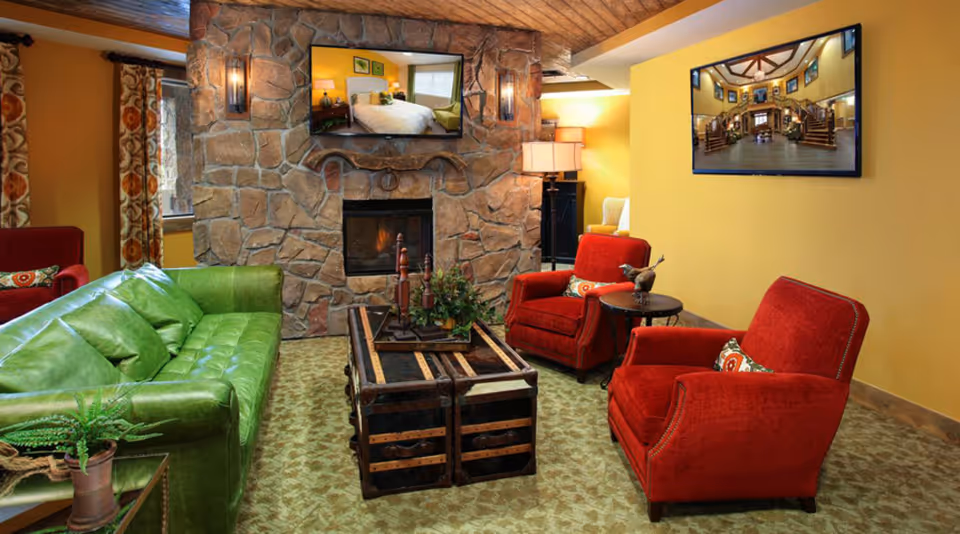 A cozy living room with a green leather sofa, two red armchairs, and a wooden trunk-style coffee table. The room features a stone fireplace with a flat-screen TV mounted above it, and another TV mounted on a yellow wall showing an image of a grand staircase. There are patterned curtains on the windows and a table lamp in the corner.