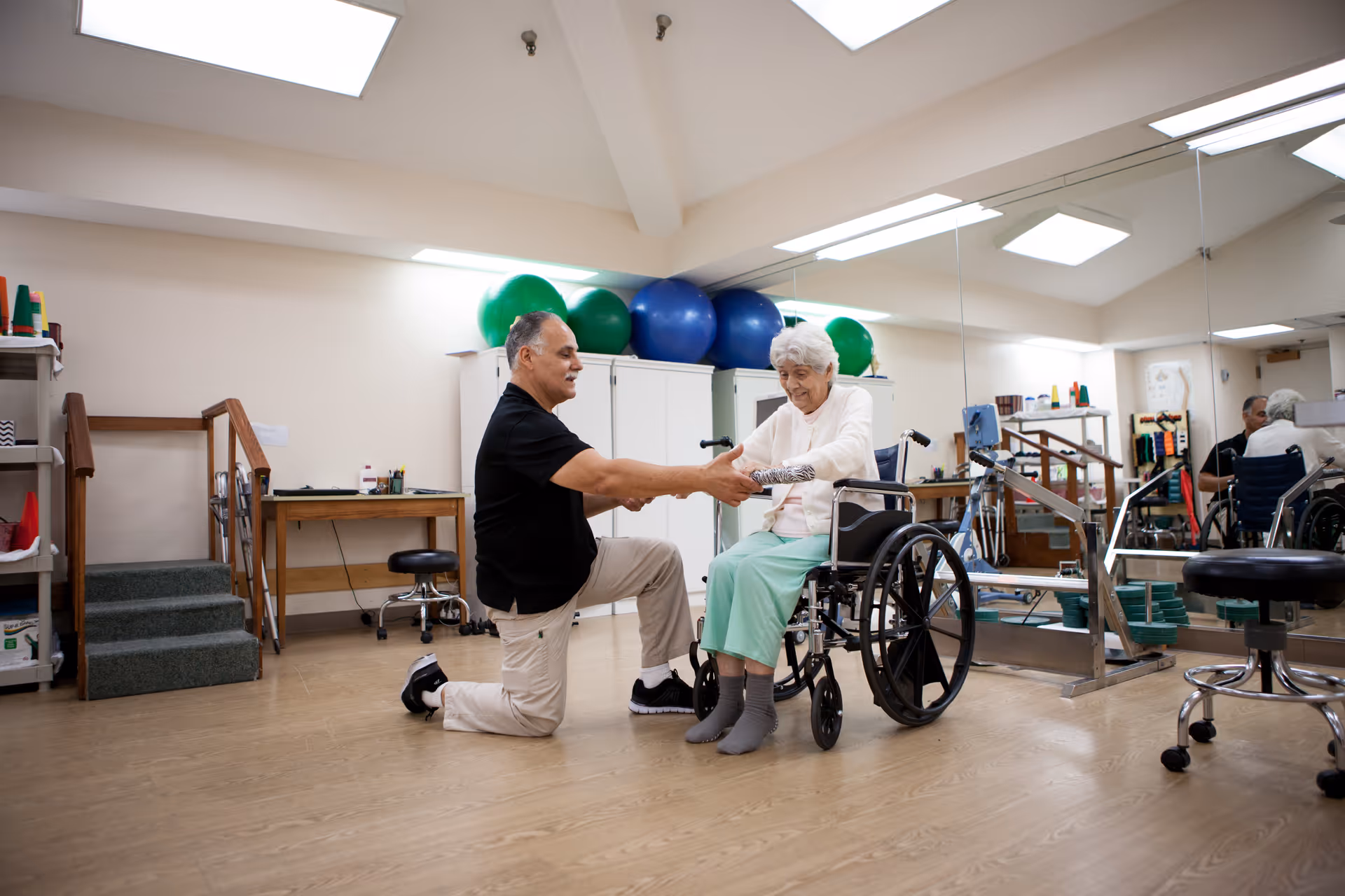 A man kneeling on one knee assists an elderly woman in a wheelchair with an arm exercise using a lightweight bar in a rehabilitation room with exercise balls, mirrors, and therapy equipment.