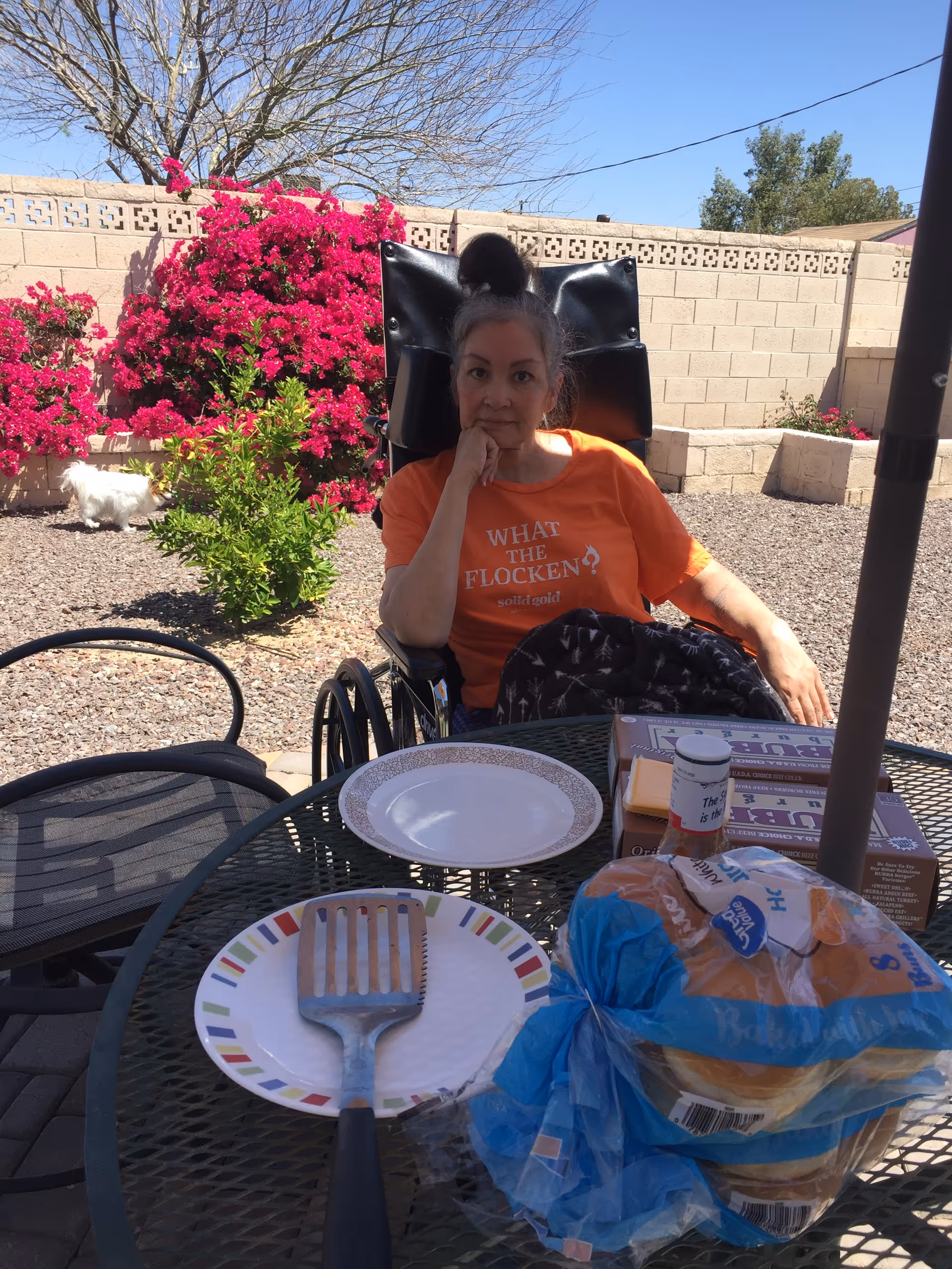 A woman in an orange shirt sitting in a wheelchair at an outdoor patio table. The table has plates, a spatula, a pack of hamburger buns, and some food containers. Behind her is a garden with bright pink flowers, a small white dog, and a brick wall under a clear blue sky.