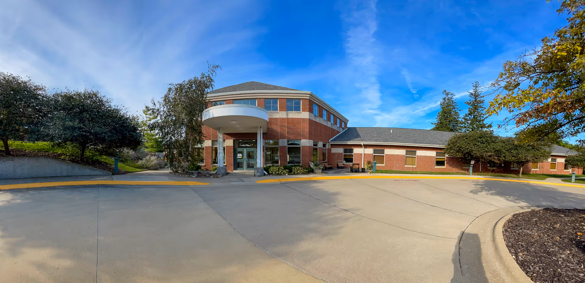 Exterior view of a two-story brick building with a circular driveway in front. The building has large windows and a covered entrance with a round canopy. Trees and shrubs surround the building under a blue sky with some clouds.