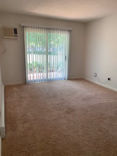 Empty room with beige carpet, white walls, a window air conditioning unit, and vertical blinds covering a sliding glass door that leads outside to a green area.