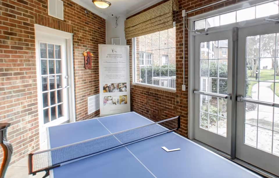 Indoor recreational area with a blue ping pong table in the center, surrounded by brick walls and large windows. A door with glass panes leads outside to a garden path. A standing banner with information about Abbotswood at Stonehenge is visible in the corner.