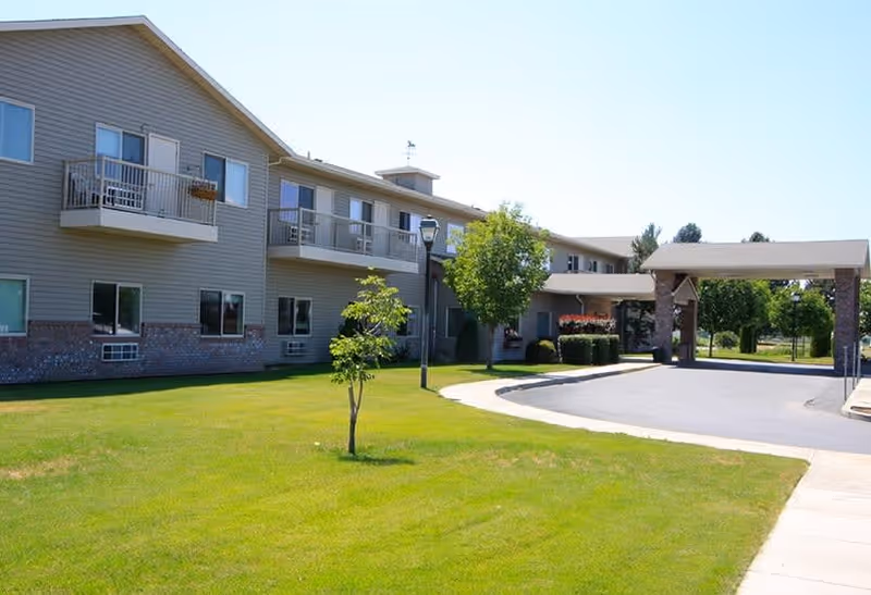Exterior view of a two-story senior living facility building with balconies, a covered entrance, a paved driveway, green lawn, and small trees under a clear blue sky.