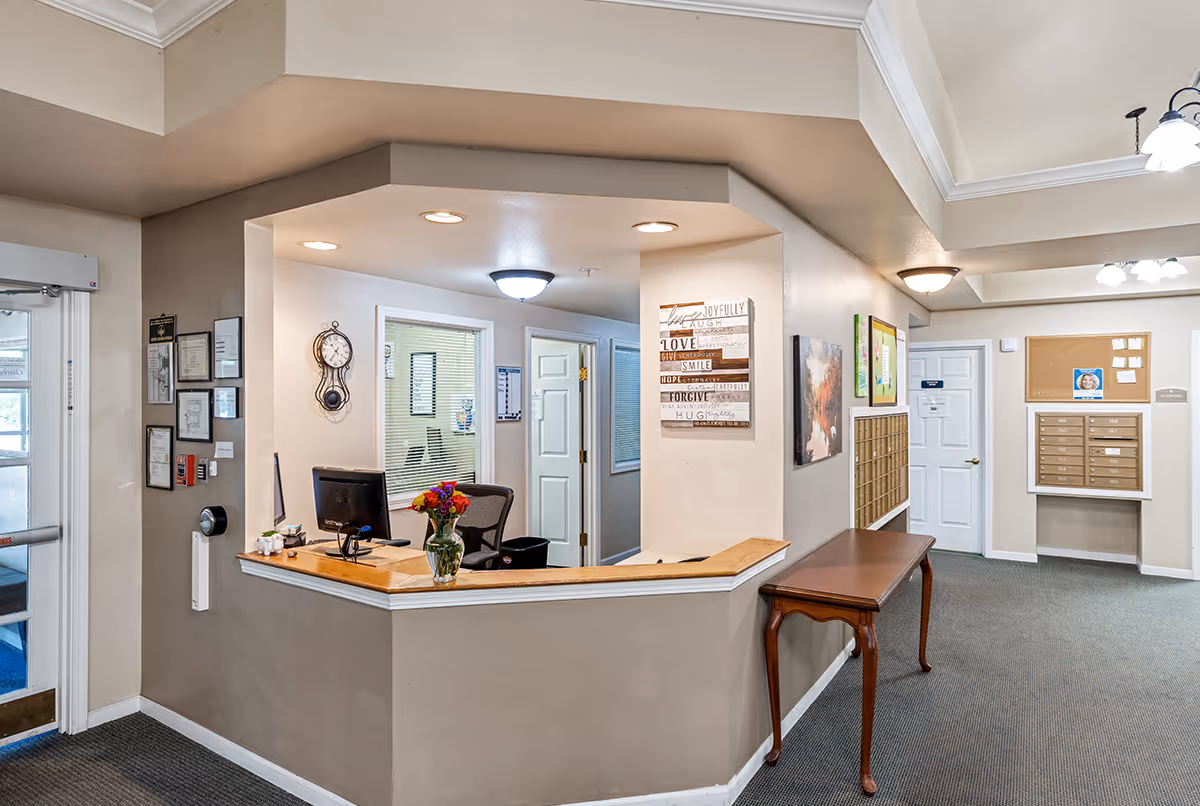 Reception area of Rolling Meadows Senior Living with a wooden desk, computer, and a vase of flowers. The walls have framed certificates, a clock, and decorative artwork. There are mailboxes and a bulletin board on the right side of the hallway.