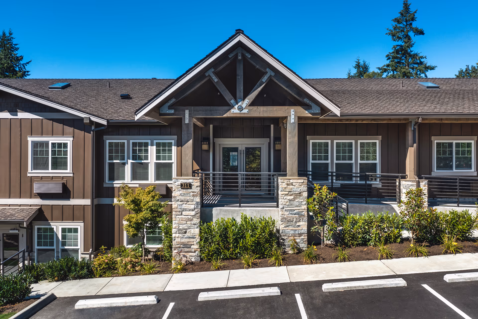 Exterior front view of a senior living facility building with brown siding, white-trimmed windows, a covered entrance supported by stone pillars, and landscaped greenery in front. The sky is clear and blue.