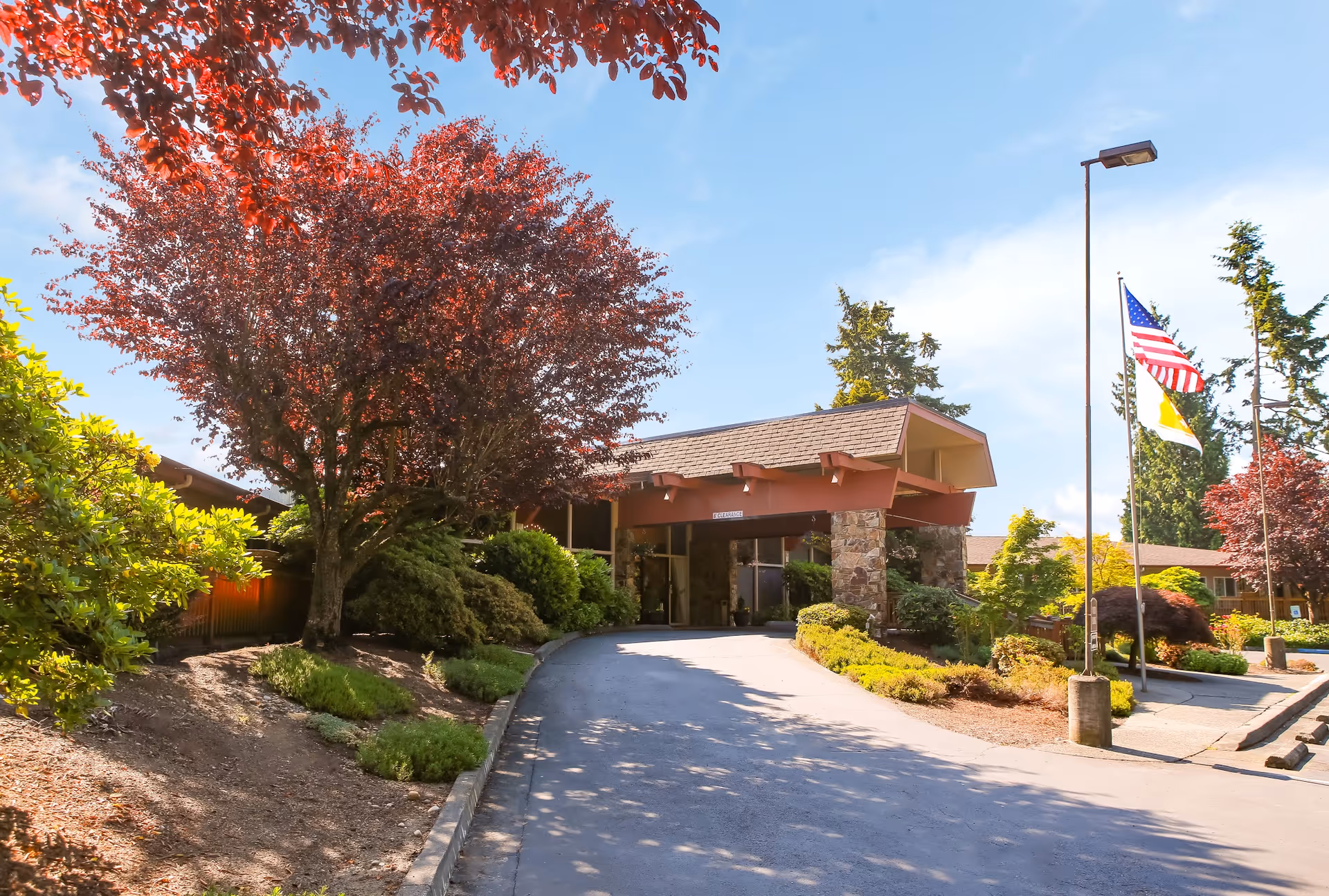 Entrance to a senior living facility with a covered driveway, surrounded by landscaped bushes and trees with red and green foliage under a clear blue sky. Two flagpoles display the American flag and another flag near the entrance.